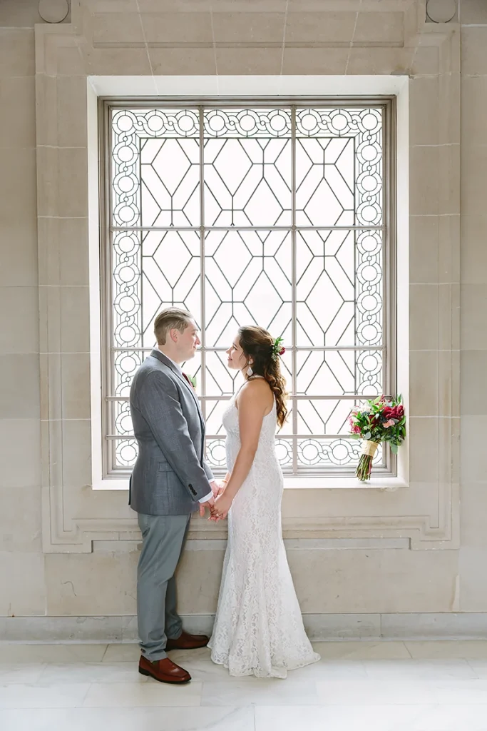 Bride and groom stand in front of window