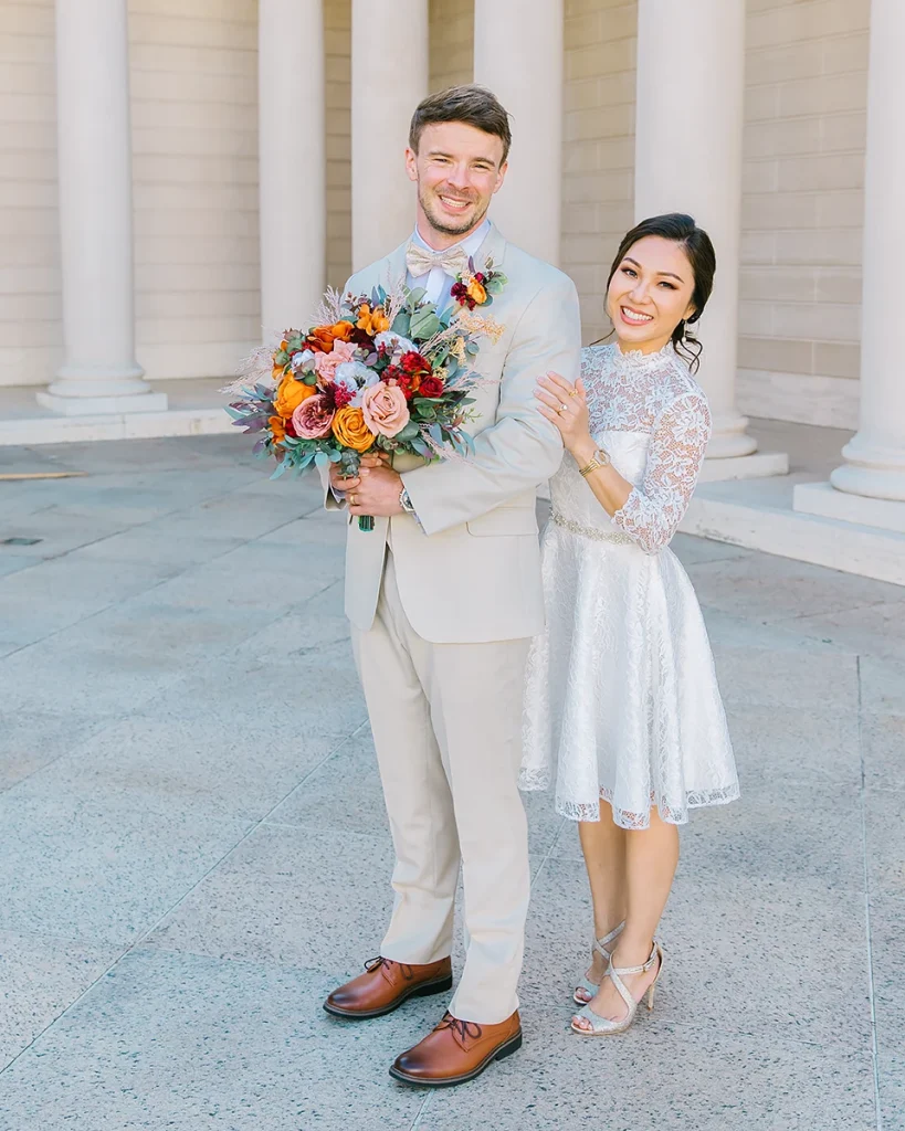 Bride stands behind groom while he holds her bridal bouqet