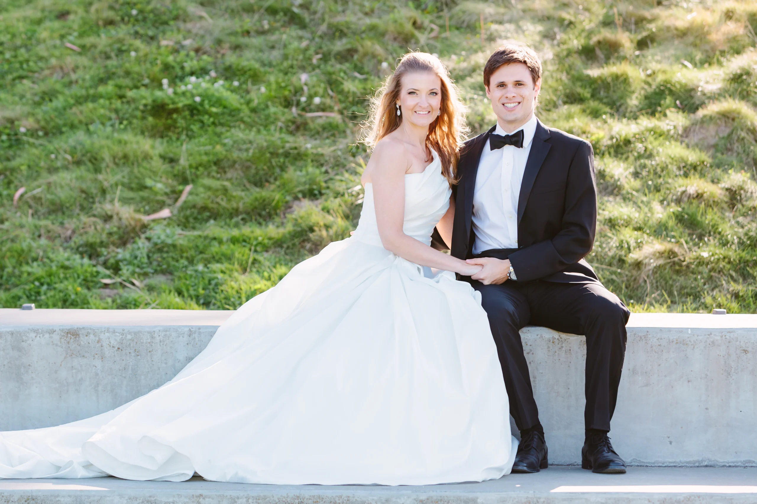 Bride and groom hold hands sitting together. Bride is wearing a gorgeous ball gown wedding dress and groom is wearing a black tuxedo